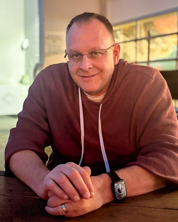 Portrait of Kipp Ransom, a man with short brown hair sitting indoors at a wooden table wearing glasses and a red textured hoodie)