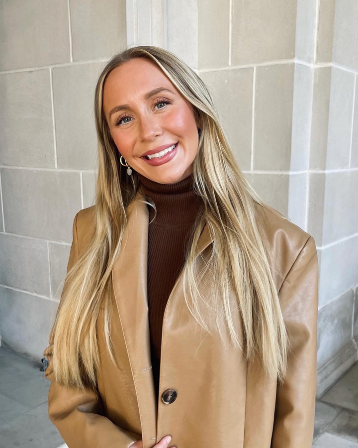 Portrait of Dr. Shaylee Schroeder (a blonde female with a brown turtleneck top and a tan leather jacket) outside in front of a tan-bricked building